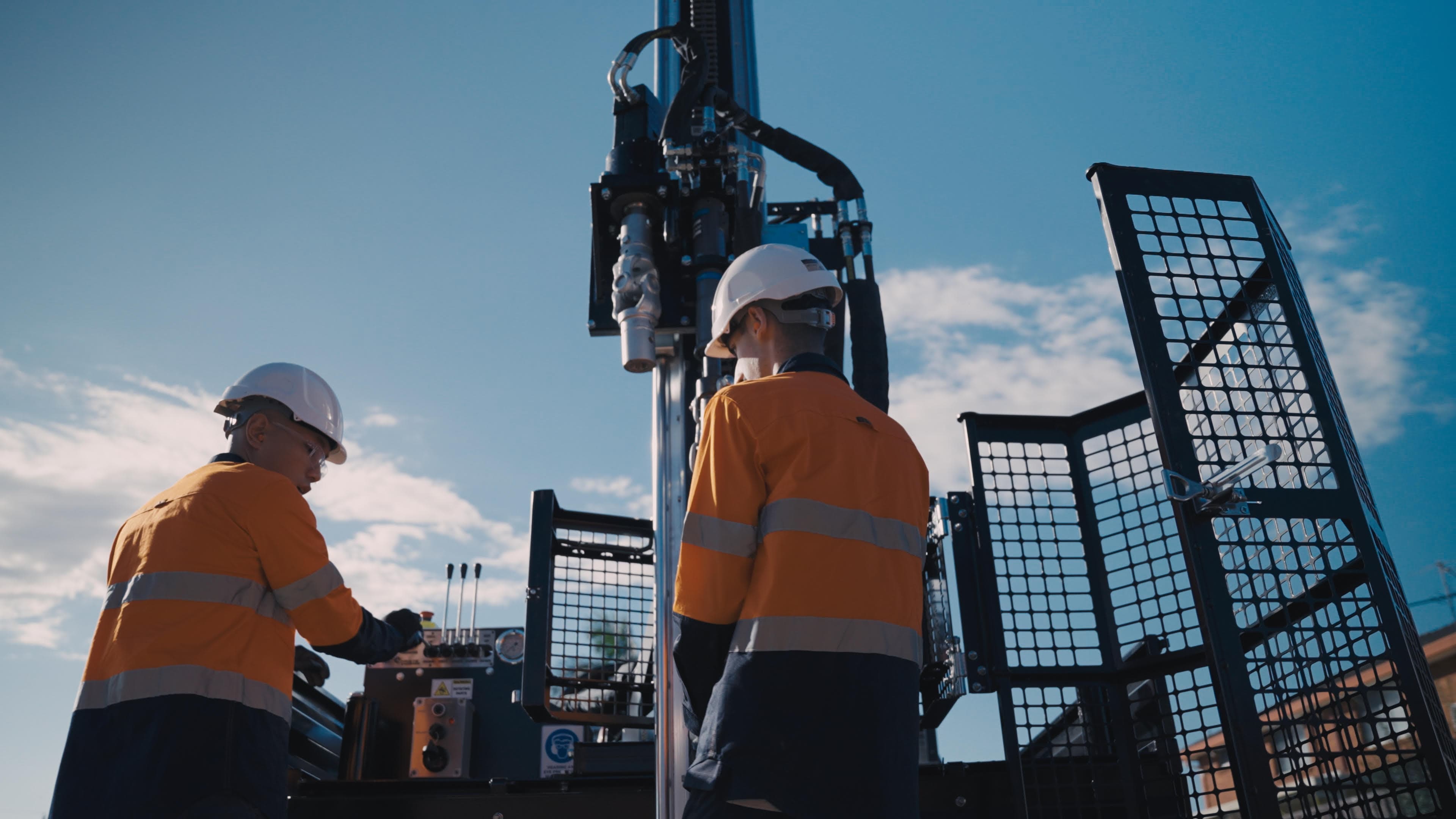 Two field engineers in high-visibility shirts and hard hats working at a drill rig against a blue sky